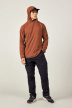 A person stands slightly turned, wearing the 686 Men's Let's Go Tech Hoody in rust for sun protection, paired with black pants and black athletic shoes with white soles, against a plain light background.