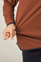 A person wearing the 686 Men's Let's Go Tech Hoody in brown and dark pants pinches and pulls the side of the shirt with their left hand to display its texture and fit. Neutral background. Brand: 686.