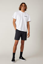 A young man with medium-length brown hair smiles with his hands in his pockets, wearing a white t-shirt, black 686 Men's Everywhere® Featherlight Chino Short, black crew socks, and black slip-on shoes against a plain light background.