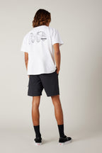 A person stands facing away, wearing a white graphic tee, 686 Men's Everywhere® Featherlight Chino Short in black, black socks, and black sneakers with white soles, set against a plain light background.