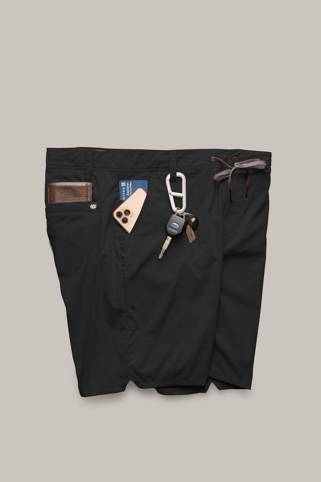 A pair of 686 Men's Everywhere® Featherlight Chino Shorts in black, with a brown leather wallet, a smartphone in a tan case, a blue access card, keys on a carabiner, and a brown braided belt tucked into the waistband and pockets.