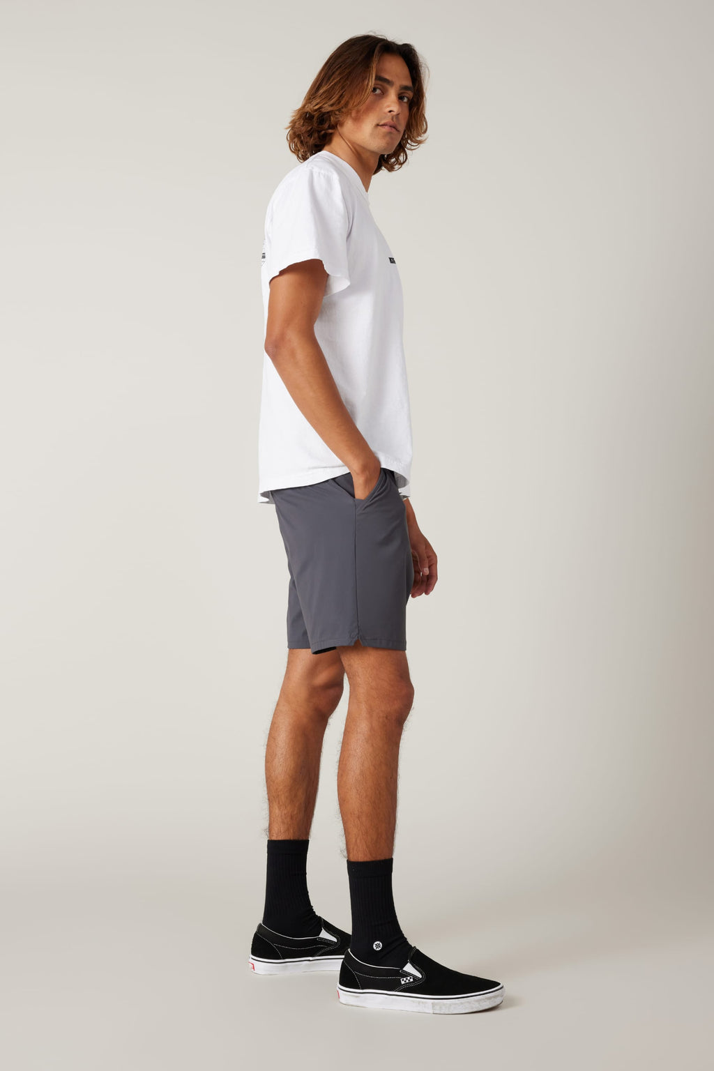 A person with shoulder-length hair stands in profile, wearing a white t-shirt, 686 Men's Everywhere® Featherlight Chino Short in gray, black crew socks, and black slip-on sneakers against a plain light background.