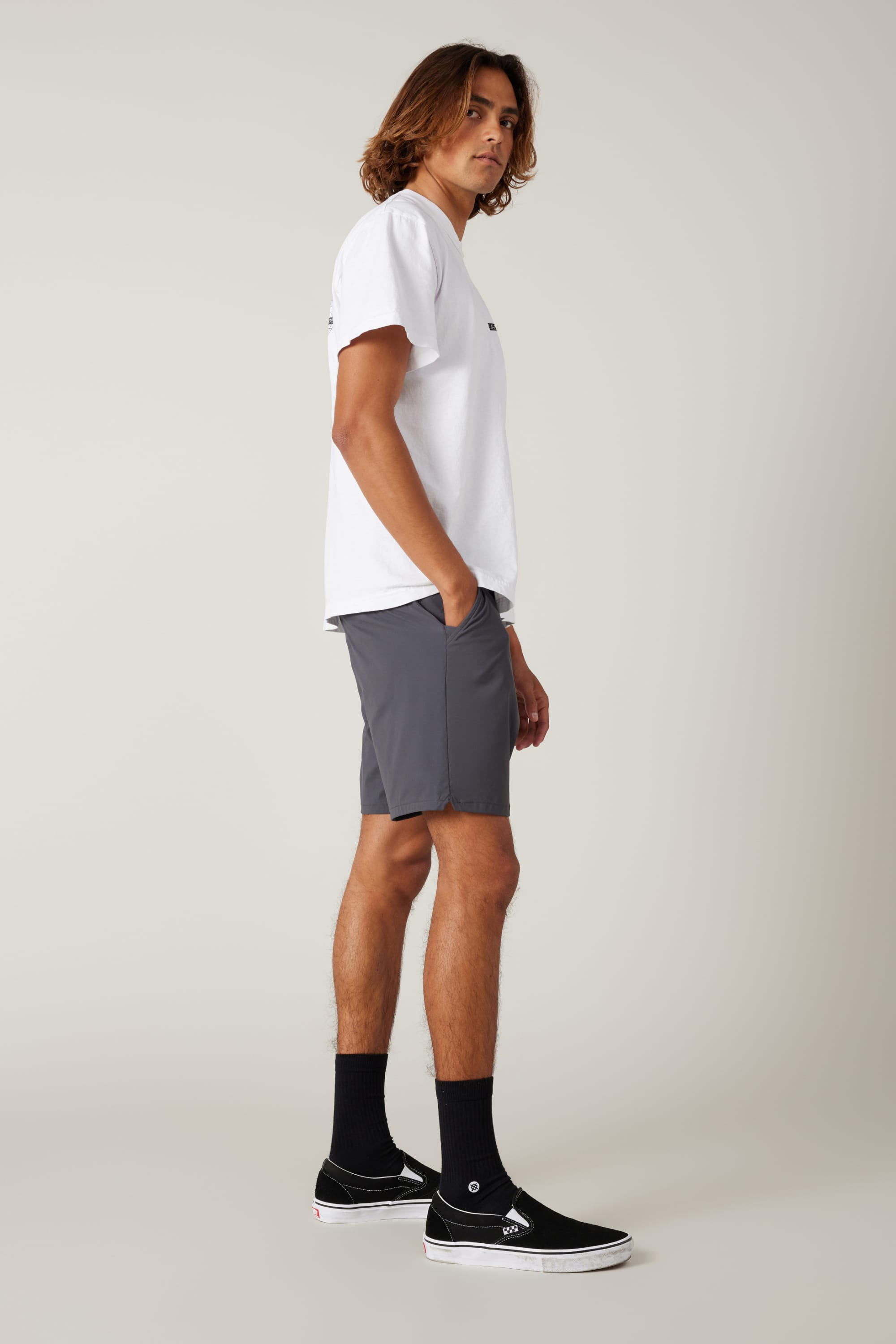 A person with shoulder-length hair stands in profile, wearing a white t-shirt, 686 Men's Everywhere® Featherlight Chino Short in gray, black crew socks, and black slip-on sneakers against a plain light background.