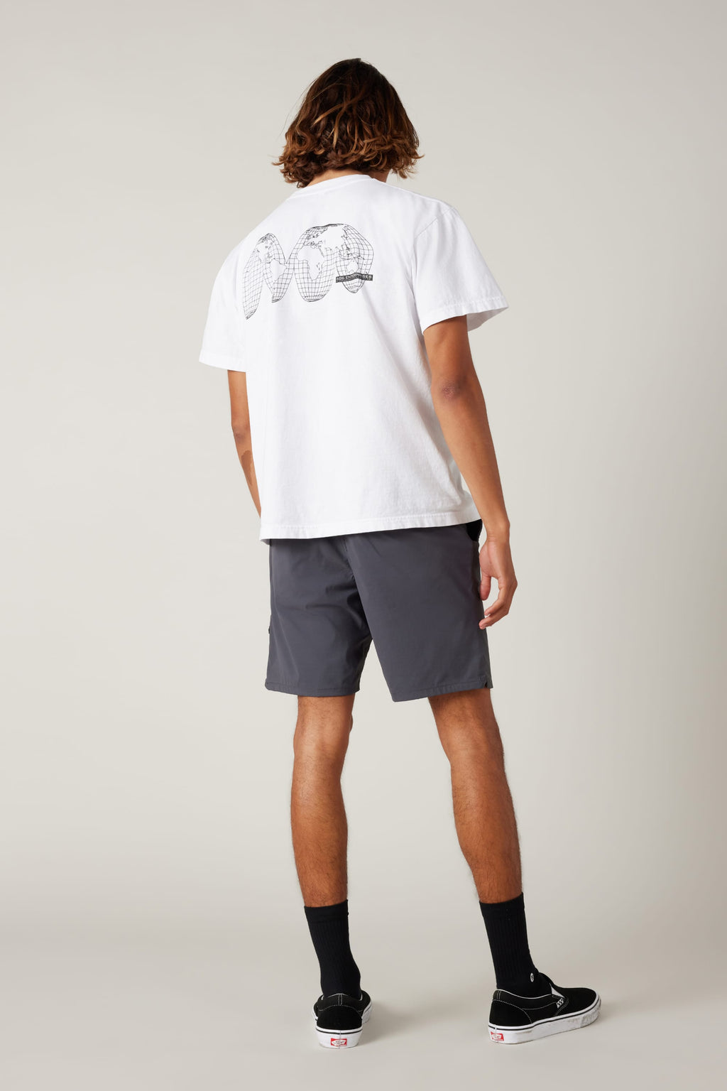 A person with shoulder-length hair wears a white graphic tee, 686 Men's Everywhere® Featherlight Chino Shorts by 686, black socks, and black sneakers. Facing away, the shirt’s back shows spherical designs on a plain light background.