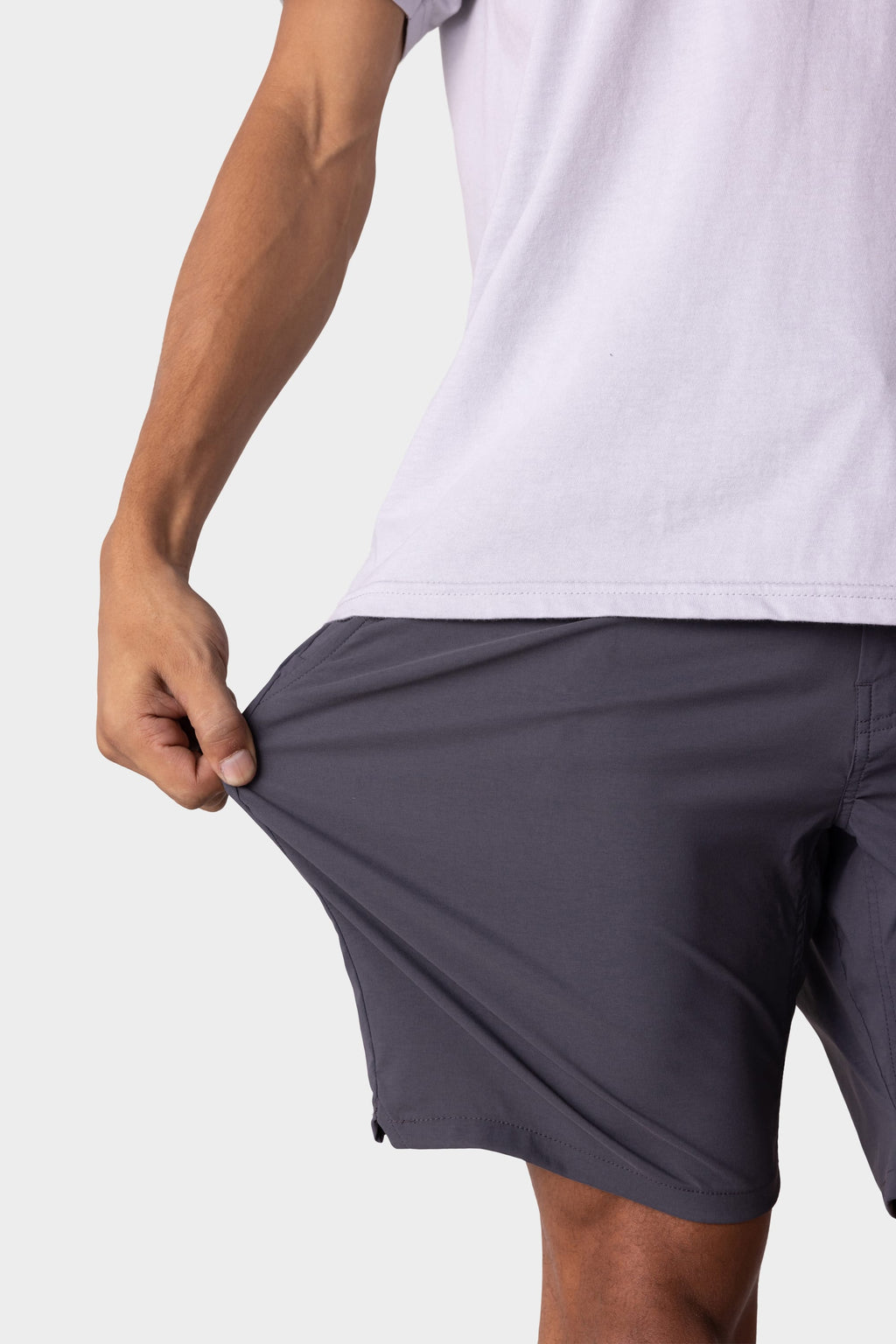 A person wearing a white T-shirt and 686 Men's Everywhere® Featherlight Chino Short in dark gray stretches the shorts' fabric outward with one hand, showcasing the flexibility of these lightweight 686 shorts.