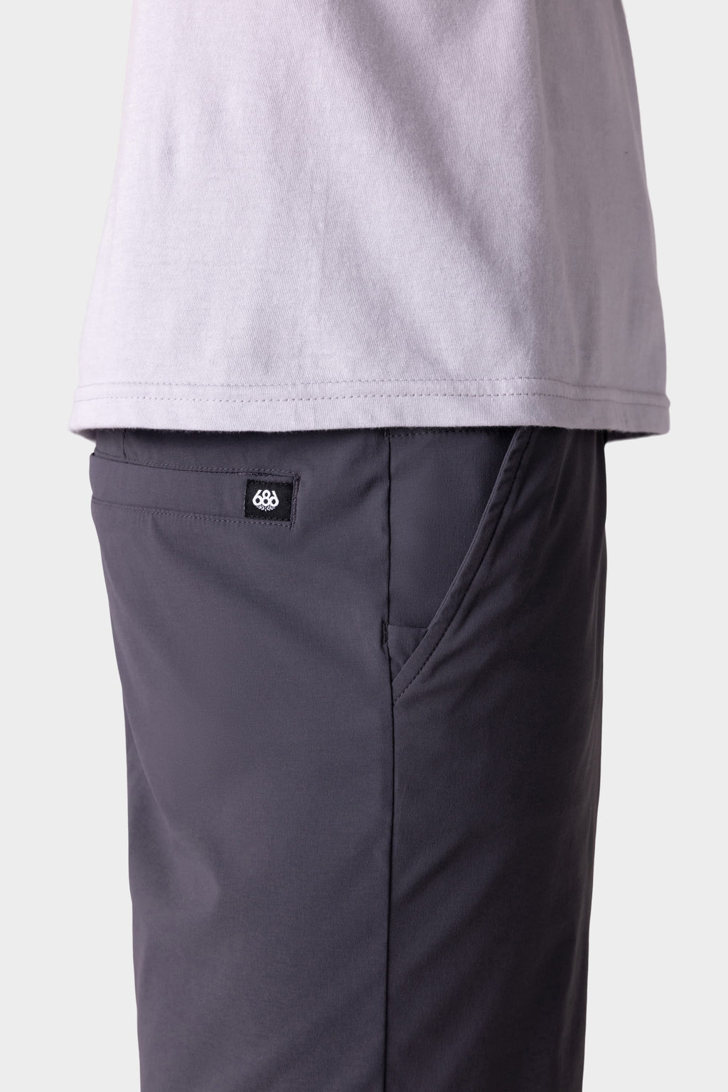 A side view shows a person wearing a light gray T-shirt and 686 Men's Everywhere® Featherlight Chino Shorts in dark gray, with a visible black and white 686 brand tag above the pocket. The background is plain and light-colored.