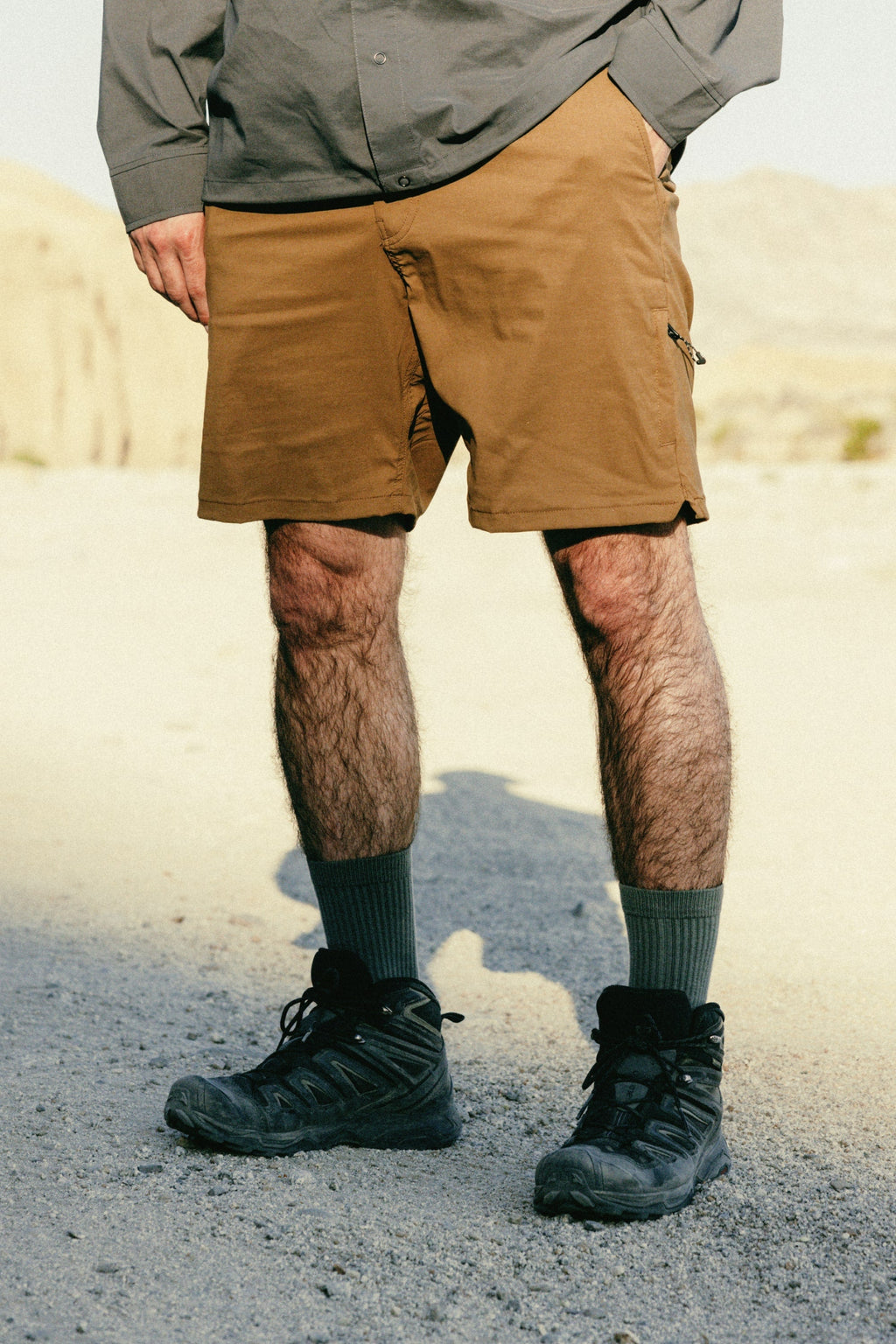 A person stands outdoors on gravel, wearing 686 Men's Everywhere® Featherlight Chino Shorts in brown, a gray long-sleeve shirt, gray socks, and black hiking boots. The sunlit scene features a natural rocky background.