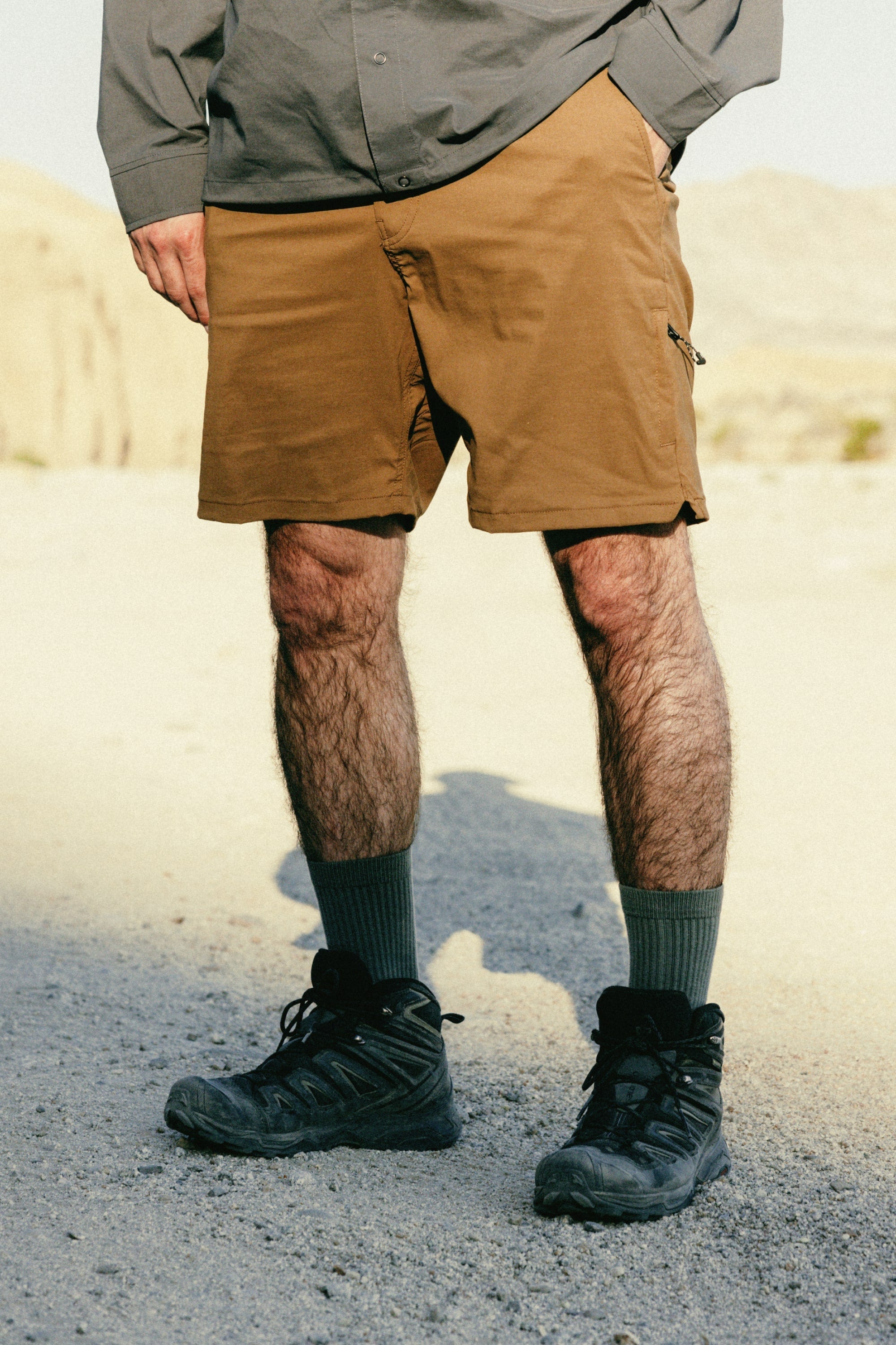 A person stands outdoors on gravel, wearing 686 Men's Everywhere® Featherlight Chino Shorts in brown, a gray long-sleeve shirt, gray socks, and black hiking boots. The sunlit scene features a natural rocky background.