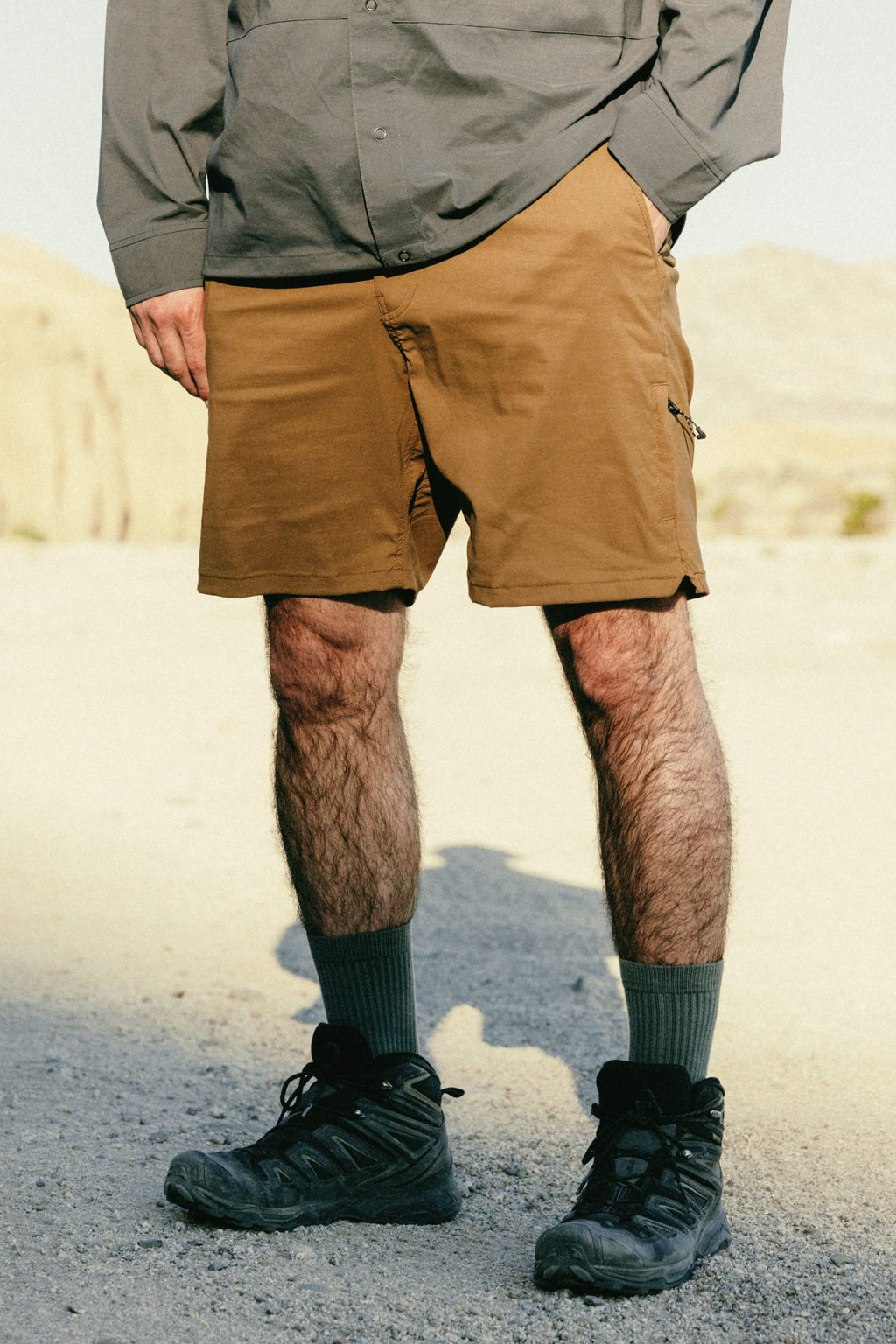 A person stands outdoors on gravel, wearing green crew socks, black hiking shoes, and 686 Men's Everywhere® Featherlight Chino Short by 686 in tan. The gray long-sleeve shirt is partly visible, with the photo cropped below the torso.