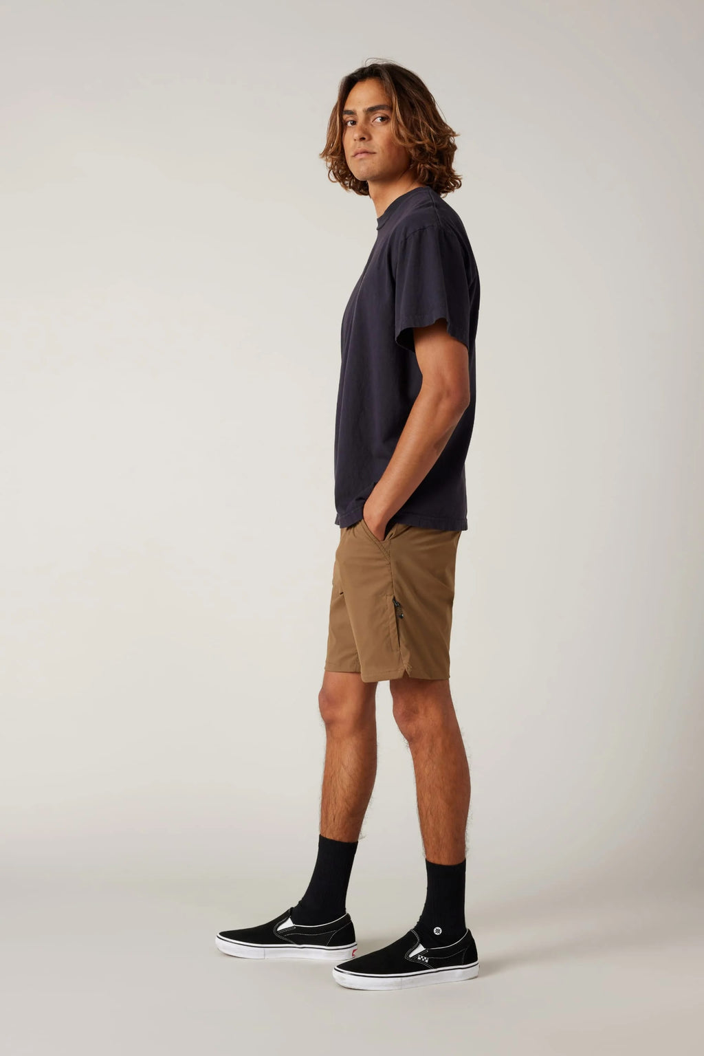 A young man with medium-length wavy hair stands in profile against a plain background, wearing a dark t-shirt, black sneakers, and 686 Men's Everywhere® Featherlight Chino Shorts—ideal for all-day comfort—with his hands in his pockets.