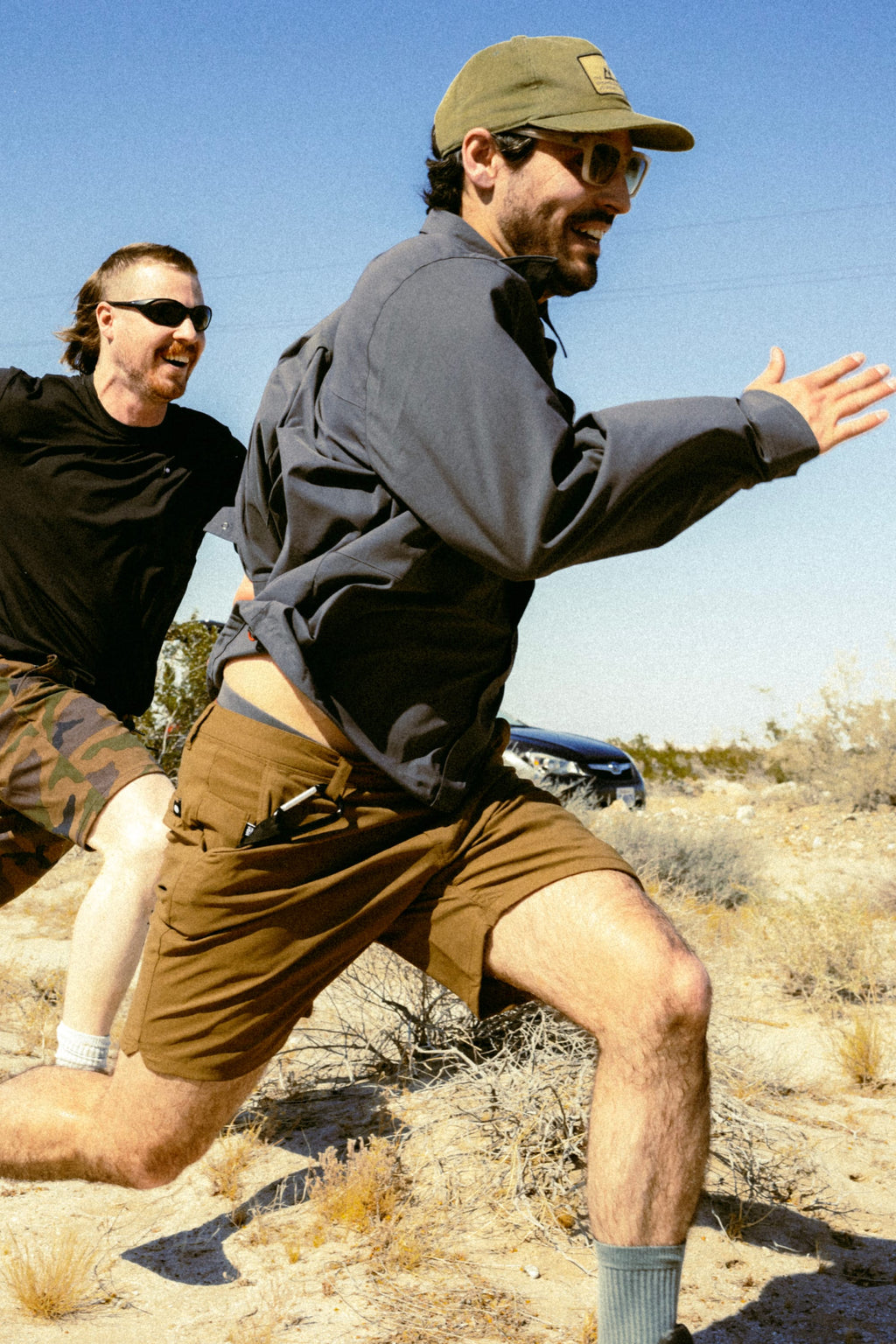Two men run energetically outdoors under a clear blue sky, wearing sunglasses, casual clothes, and 686 Men's Everywhere® Featherlight Chino Shorts by 686. The desert-like landscape features shrubs and dry grass in the background.
