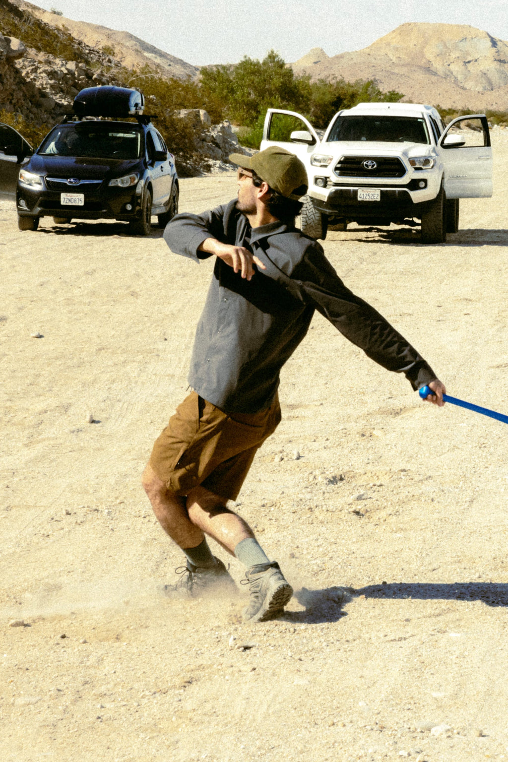 A person in 686 Men's Everywhere® Featherlight Chino Shorts, a cap, sunglasses, and a jacket swings a blue bat on dusty ground with cars and mountains in the background.