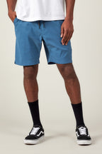 A person wearing a white T-shirt, 686 Men's Everywhere® Featherlight Chino Short by 686 in blue, black crew socks, and black Vans sneakers stands against a plain light background. The image is cropped from the waist down.