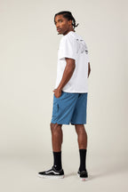 A man stands sideways, looking over his shoulder. He wears a white graphic tee, 686 Men's Everywhere® Featherlight Chino Short in blue, black crew socks, and black Vans sneakers against a plain light background.