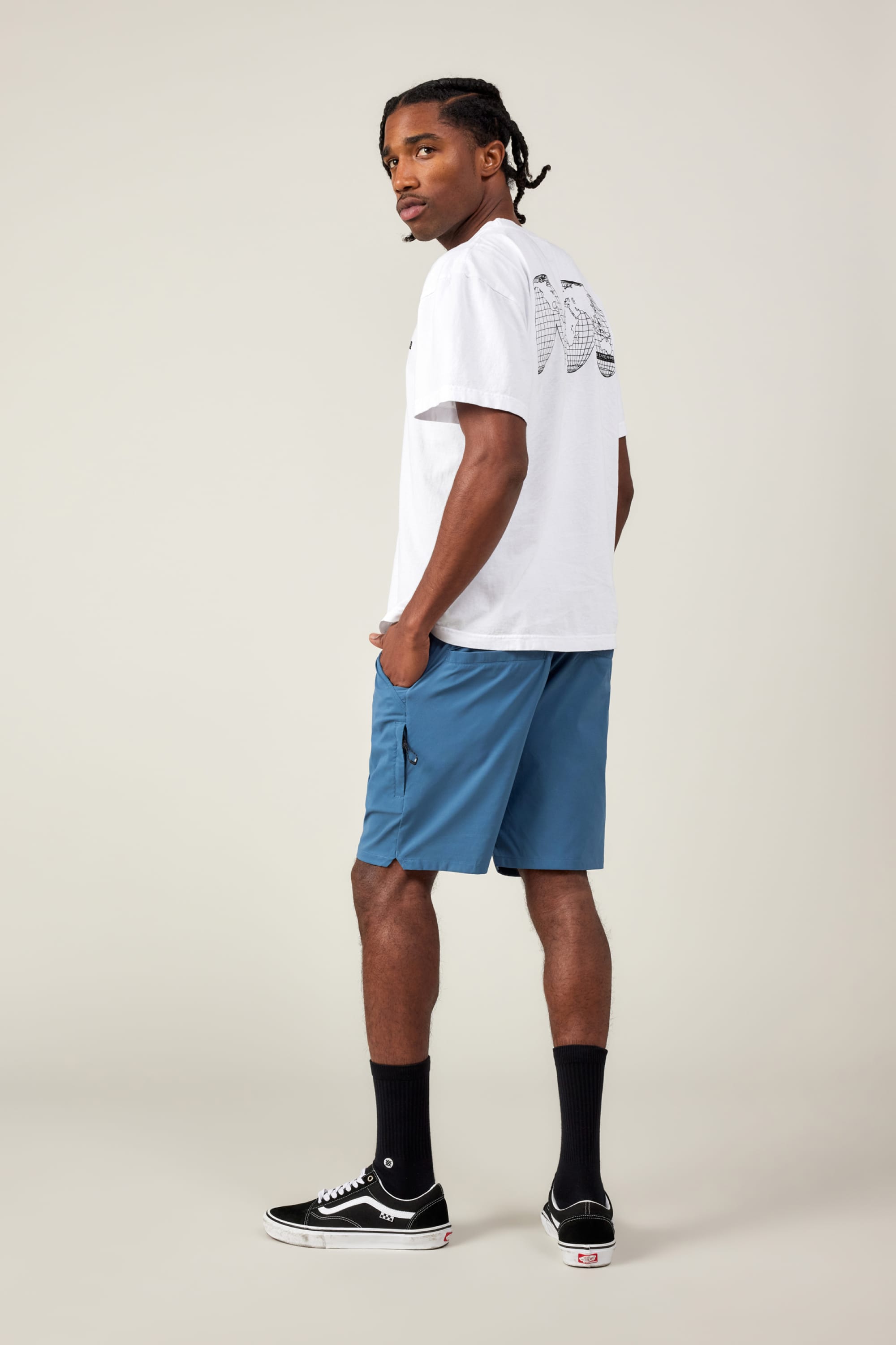 A man stands sideways, looking over his shoulder. He wears a white graphic tee, 686 Men's Everywhere® Featherlight Chino Short in blue, black crew socks, and black Vans sneakers against a plain light background.