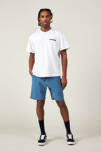 A man stands against a plain light background wearing a white t-shirt, black crew socks, black-and-white sneakers, and 686 Men's Everywhere® Featherlight Chino Short in blue by 686. He faces forward, hands in pockets, with a neutral expression.