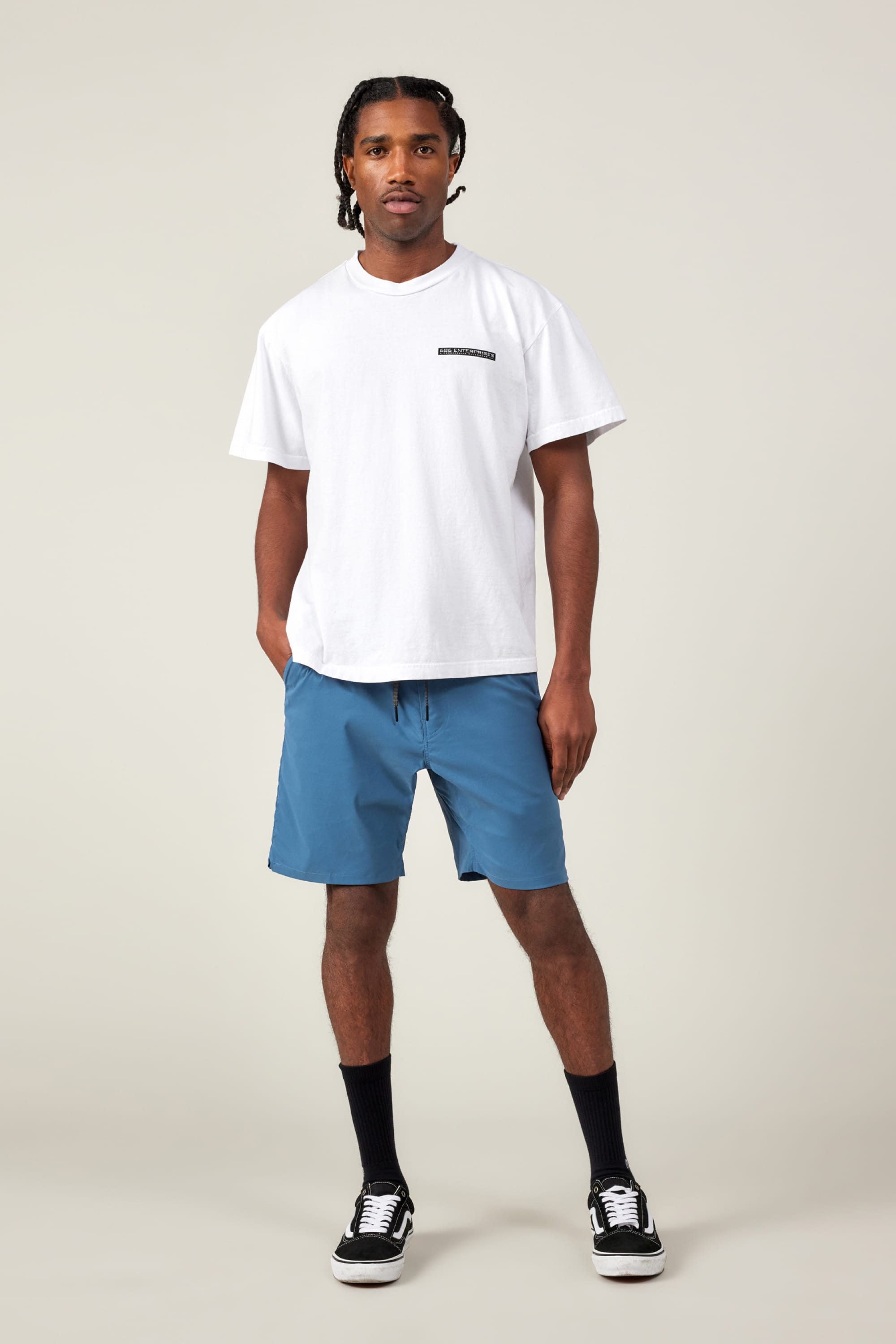 A man stands against a plain light background wearing a white t-shirt, black crew socks, black-and-white sneakers, and 686 Men's Everywhere® Featherlight Chino Short in blue by 686. He faces forward, hands in pockets, with a neutral expression.