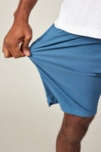 A person wearing a white shirt and 686 Men's Everywhere® Featherlight Chino Shorts in blue stretches the fabric outward with one hand, demonstrating the flexibility of these 686 shorts.