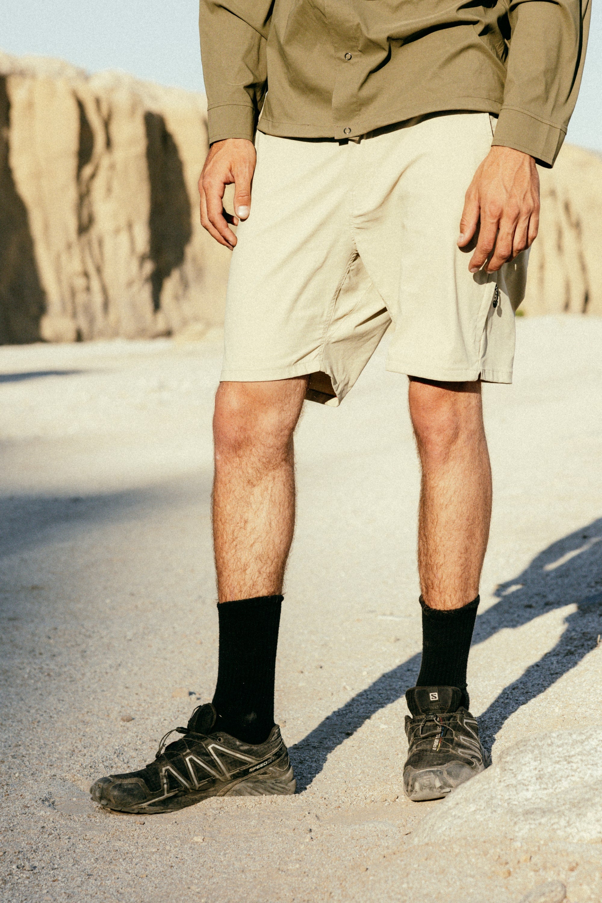 On rocky terrain outdoors, a person wears 686 Men's Everywhere® Featherlight Chino Short by 686, paired with a green long-sleeve shirt, black socks, and black hiking shoes. Their head and upper torso are not shown in the image.