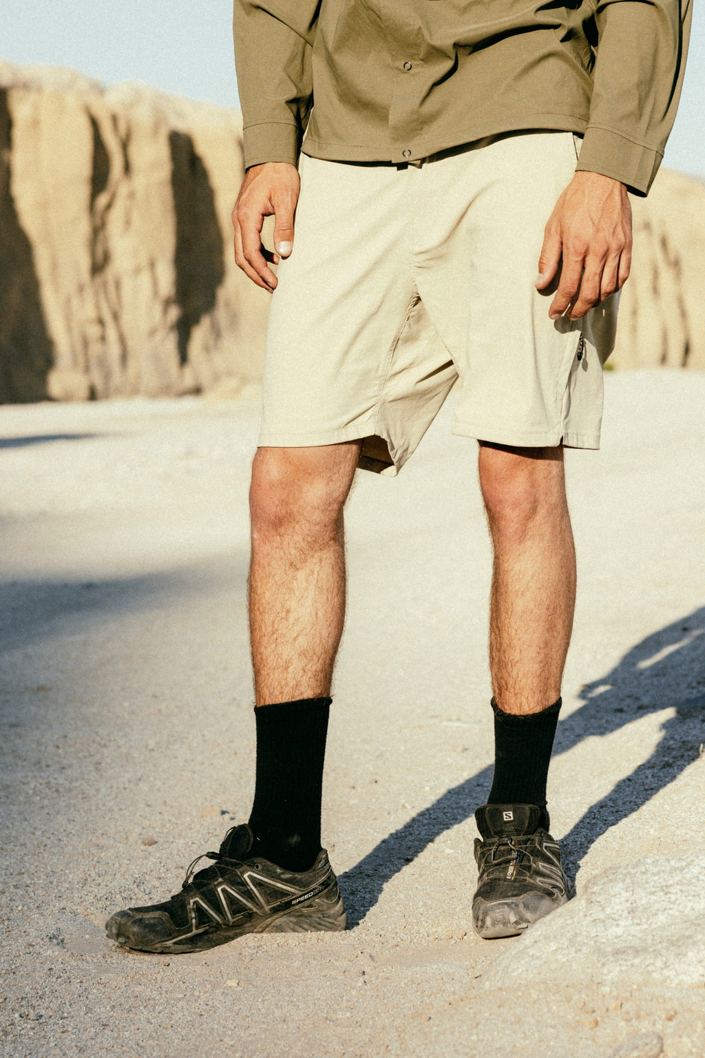A person stands on a sunlit rocky terrain with cliffs in the background, wearing 686 Men's Everywhere® Featherlight Chino Short, a green shirt, black socks, and black trail shoes. The photo is cropped above the shoulders.