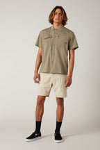 A man with medium-length brown hair and a relaxed posture wears black slip-on sneakers, black socks, an olive-green button-up shirt, and 686 Men's Everywhere® Featherlight Chino Shorts by 686, standing against a plain background.