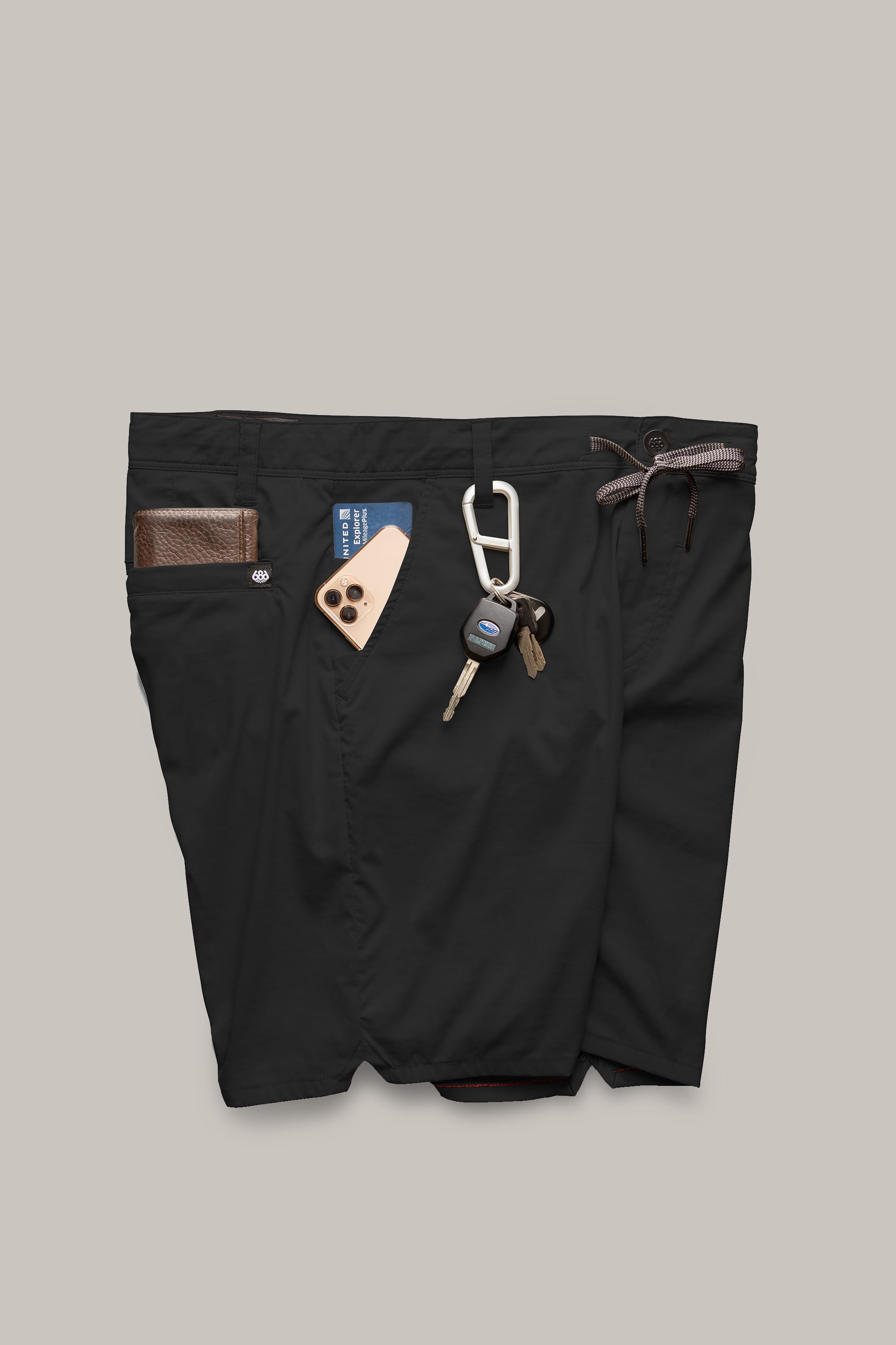 A pair of 686 Men's Everywhere® Featherlight Chino Shorts in black holds a brown wallet, smartphone, keys on a carabiner, and a card in its waistband and pockets against a light gray background.