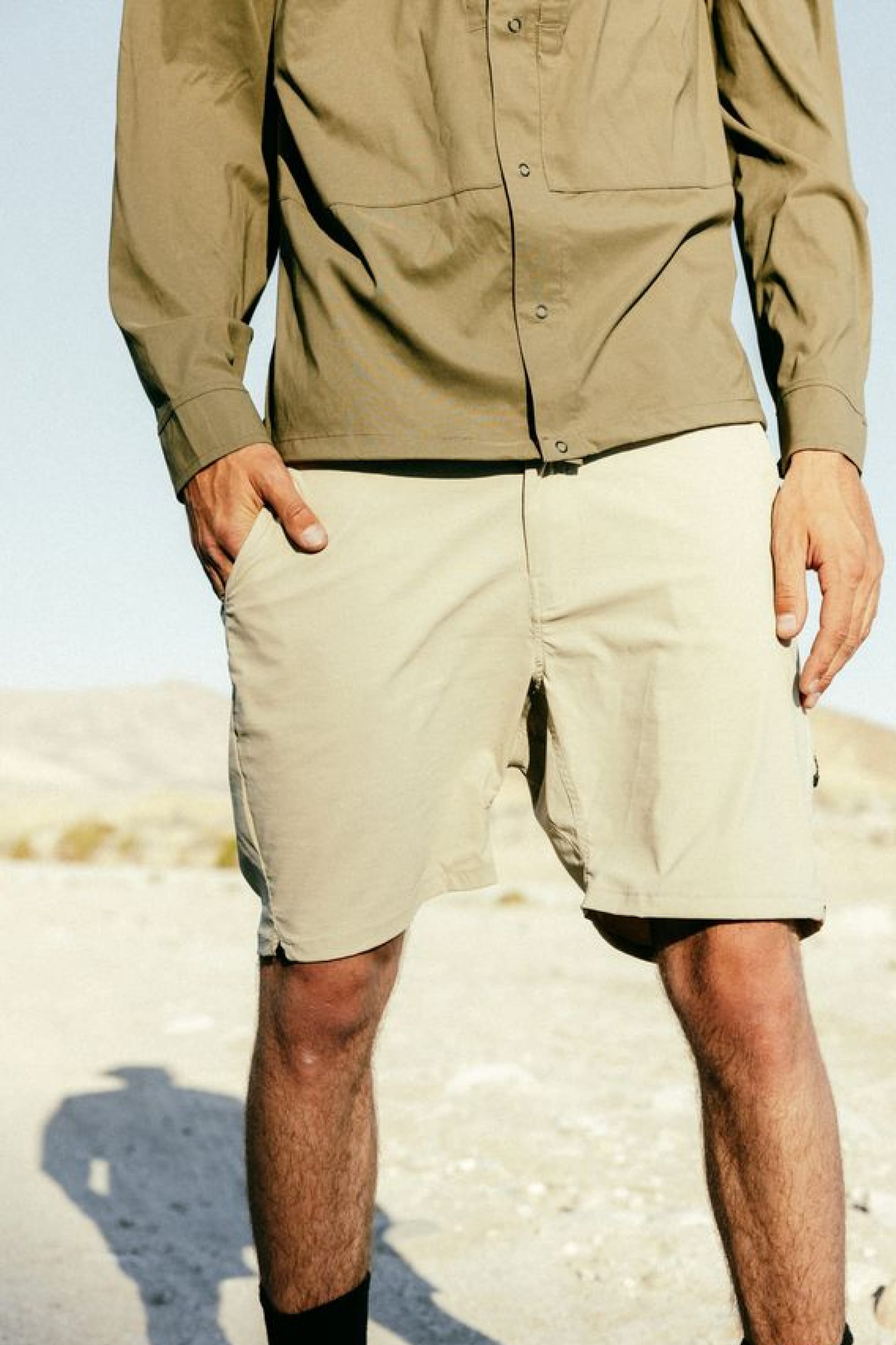 A person stands outdoors on sunlit sand, wearing a long-sleeve olive-green shirt and 686 Men's Everywhere® Featherlight Chino Short by 686. The desert landscape features distant hills in the background.