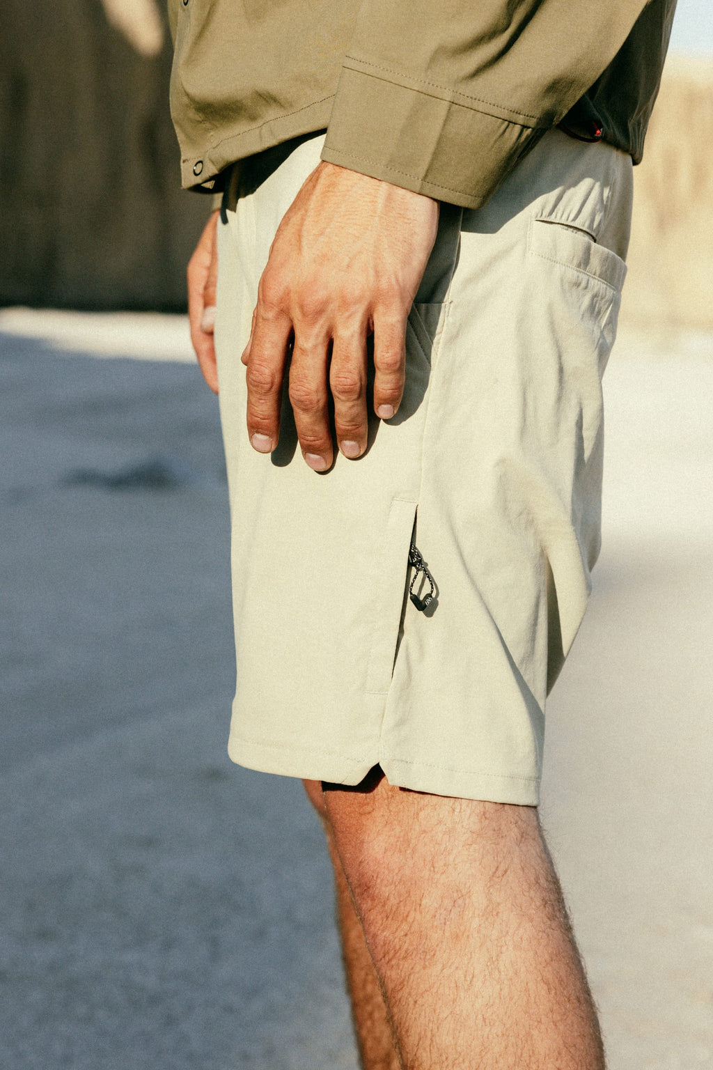 A person stands outdoors in a light green long-sleeve shirt and 686 Men's Everywhere® Featherlight Chino Short, their hand at their side highlighting the lightweight texture of the shorts in natural light.
