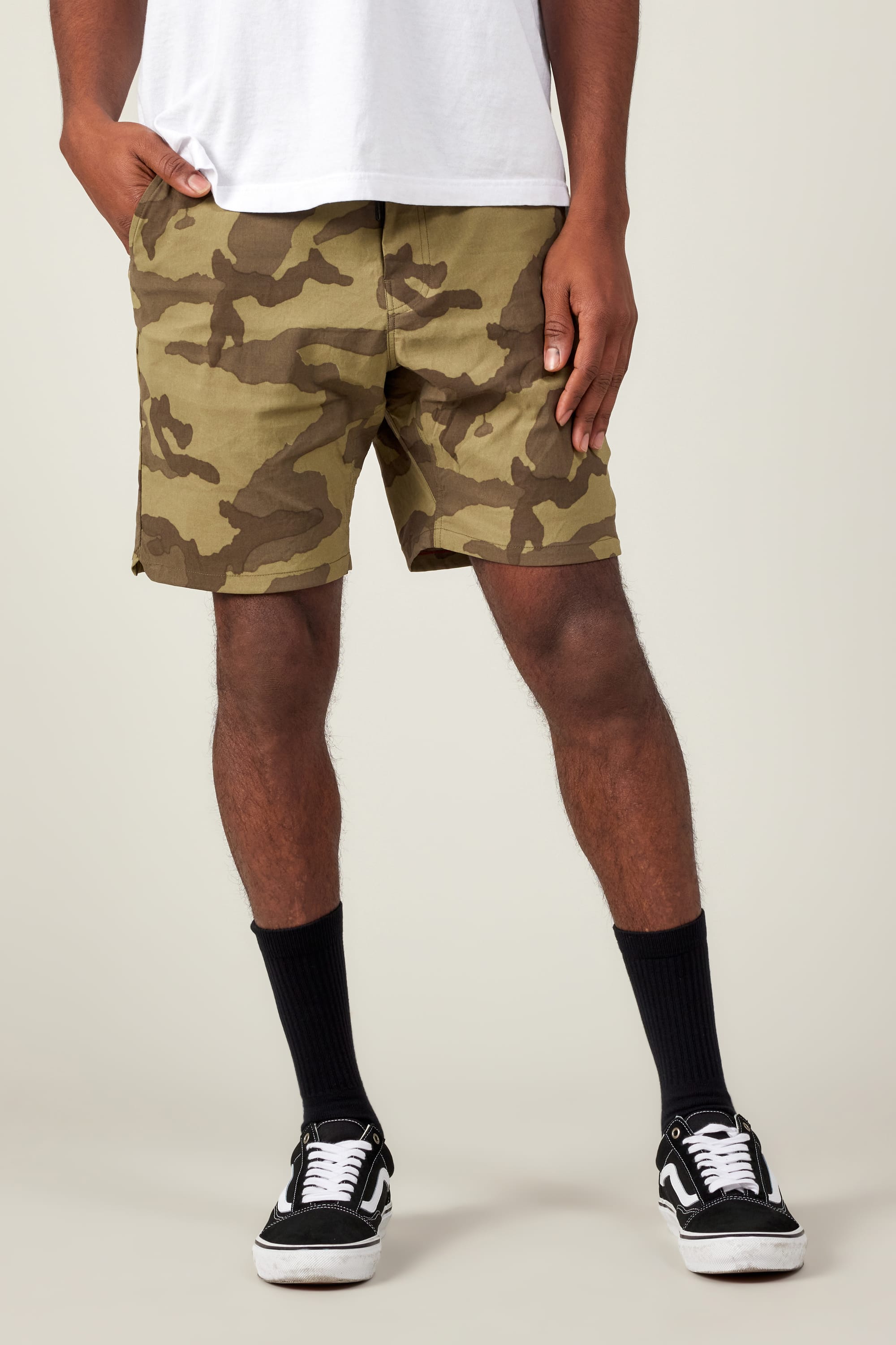 Wearing 686 Men's Everywhere® Featherlight Chino Shorts, a white T-shirt, black crew socks, and black-and-white sneakers, the person stands with one hand in their pocket against a plain background.