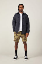 A man with short braided hair stands facing forward against a plain light background, wearing a black quilted jacket, a white t-shirt, 686 Men's Everywhere® Featherlight Chino Short by 686, black socks, and black-and-white sneakers.