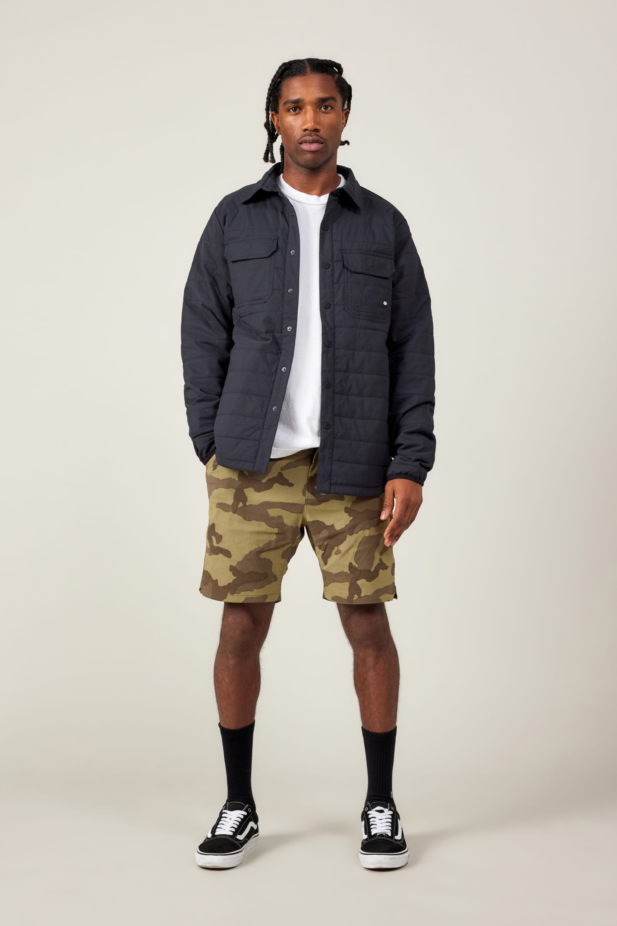 A man with short braided hair stands facing forward against a plain light background, wearing a black quilted jacket, a white t-shirt, 686 Men's Everywhere® Featherlight Chino Short by 686, black socks, and black-and-white sneakers.