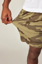 A hand pulls at the hem of 686 Men's Everywhere® Featherlight Chino Short, highlighting the stretch of its green and brown camouflage fabric. Only part of the hand, shorts, and leg are visible against a plain background.