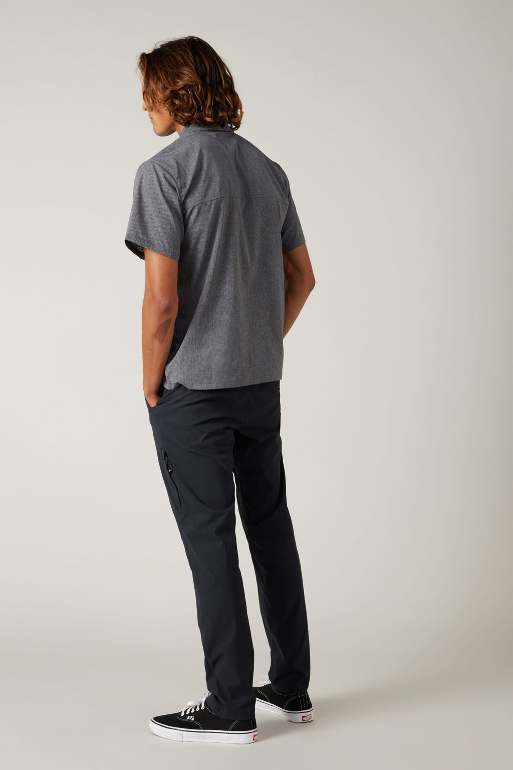 A person with shoulder-length brown hair, in a short-sleeve gray button-up shirt, 686 Men's Everywhere® Featherlight Chino Pant - Slim Fit by 686, and black sneakers stands hands-in-pockets against a plain light background. Ideal for summer.