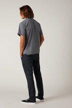 A person with shoulder-length brown hair, in a short-sleeve gray button-up shirt, 686 Men's Everywhere® Featherlight Chino Pant - Slim Fit by 686, and black sneakers stands hands-in-pockets against a plain light background. Ideal for summer.