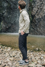 A man in a beige long-sleeve shirt, 686 Men's Everywhere® Featherlight Chino Pant - Slim Fit, and black sneakers stands on rocky ground by a waterfall—perfect inspiration for your summer wardrobe.
