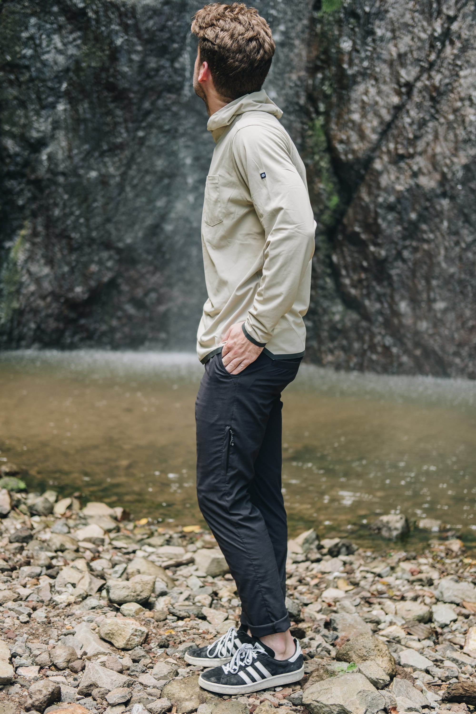 A man in a beige long-sleeve shirt, 686 Men's Everywhere® Featherlight Chino Pant - Slim Fit, and black sneakers stands on rocky ground by a waterfall—perfect inspiration for your summer wardrobe.