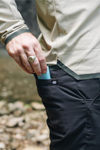 Wearing the 686 Men's Everywhere® Featherlight Chino Pant - Slim Fit in black, a person in a beige long-sleeve shirt slips a blue rectangular item into its small zippered pocket. Their silver ring shows as rocks and water blur in the background—ideal for summer.