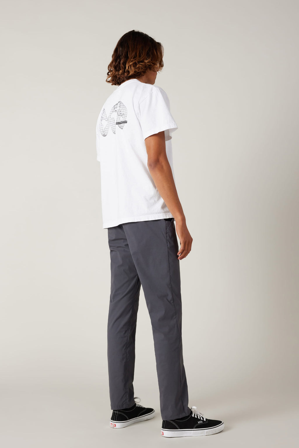 Against a plain light background, a person is seen from behind wearing a white T-shirt with a black geometric back design, 686 Men's Everywhere® Featherlight Chino Pant - Slim Fit in gray, and black Vans sneakers—ideal for summer style.