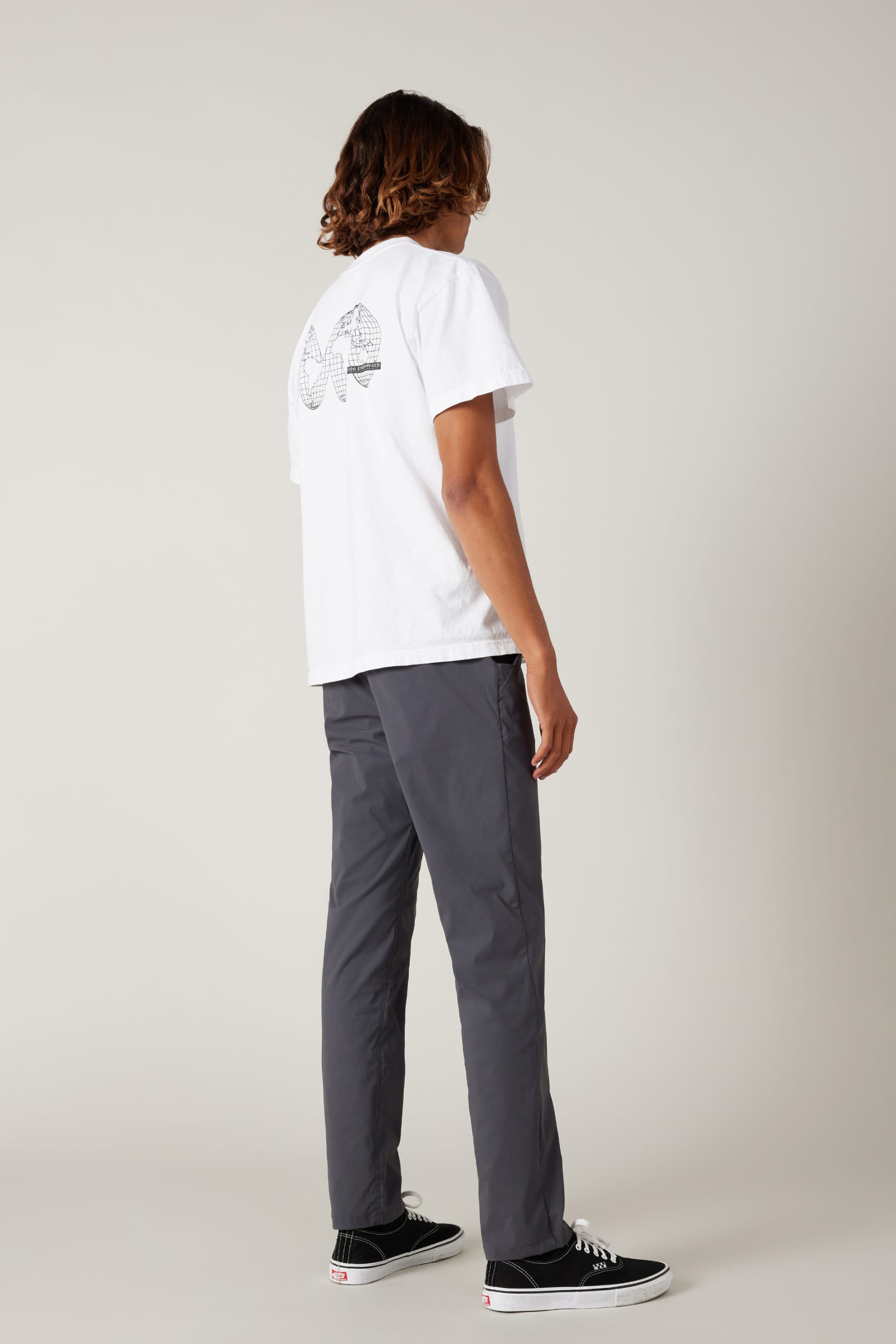 Against a plain light background, a person is seen from behind wearing a white T-shirt with a black geometric back design, 686 Men's Everywhere® Featherlight Chino Pant - Slim Fit in gray, and black Vans sneakers—ideal for summer style.