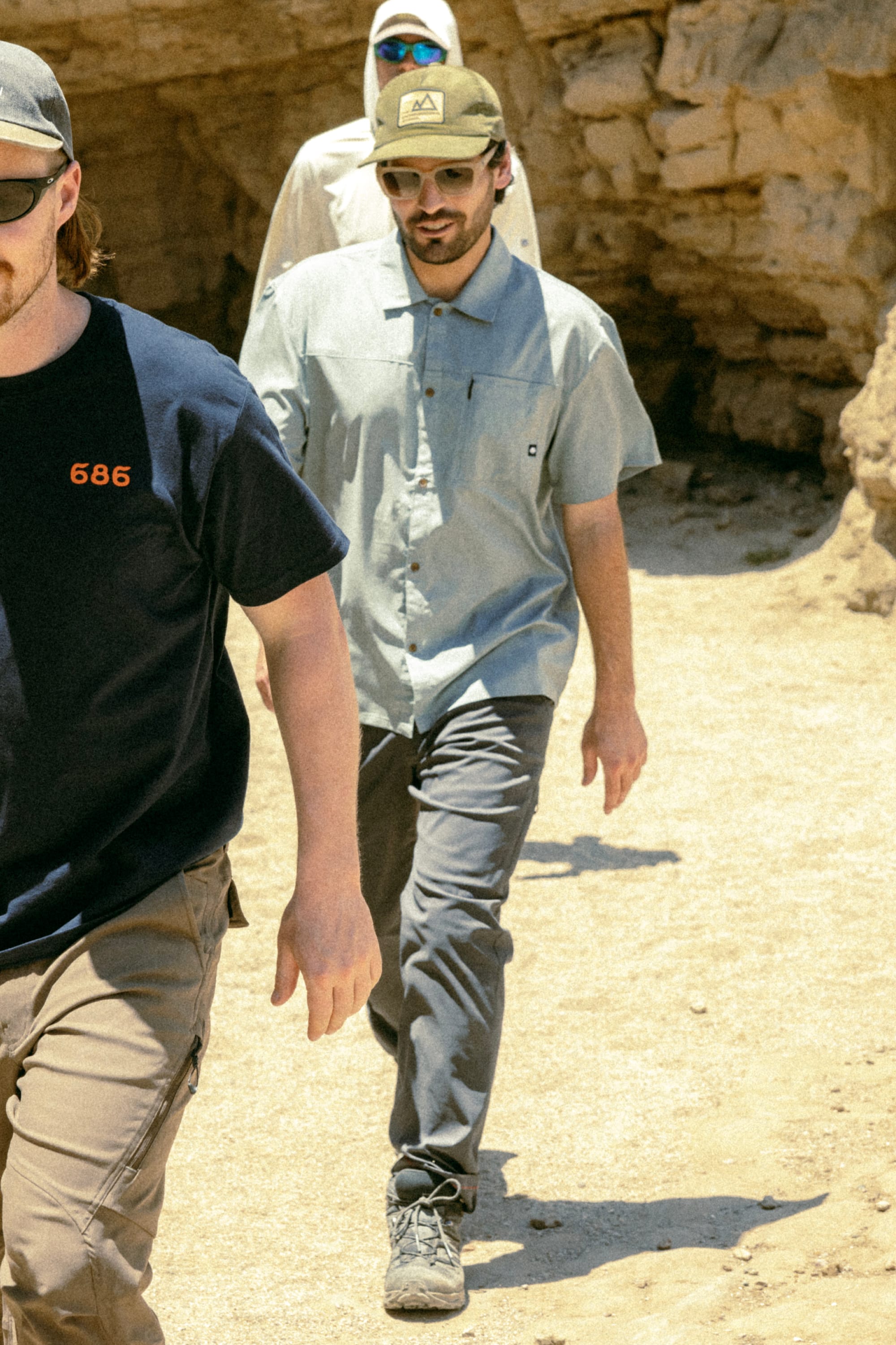 Three men walk outdoors on a sunny day by rocky terrain. Two wear hats and casual summer outfits, including the 686 Men's Everywhere® Featherlight Chino Pant - Slim Fit, ideal for staying cool and comfortable in the sun.