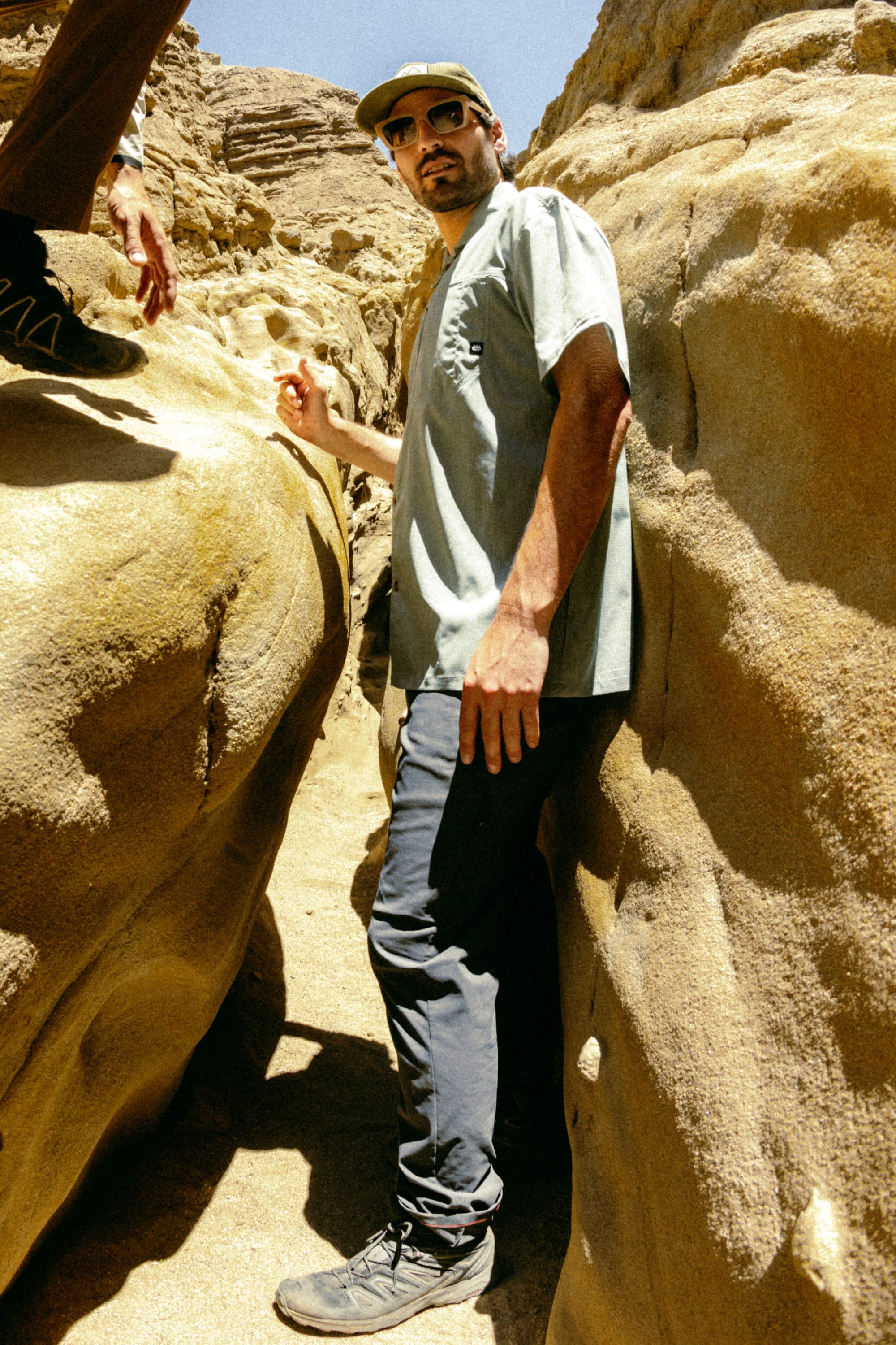 Amid sunlit canyon rocks, a man in sunglasses, a hat, and the 686 Men's Everywhere® Featherlight Chino Pant (Slim Fit) enjoys a summer day; another person’s legs and hand appear nearby, showcasing a perfect warm-weather wardrobe.