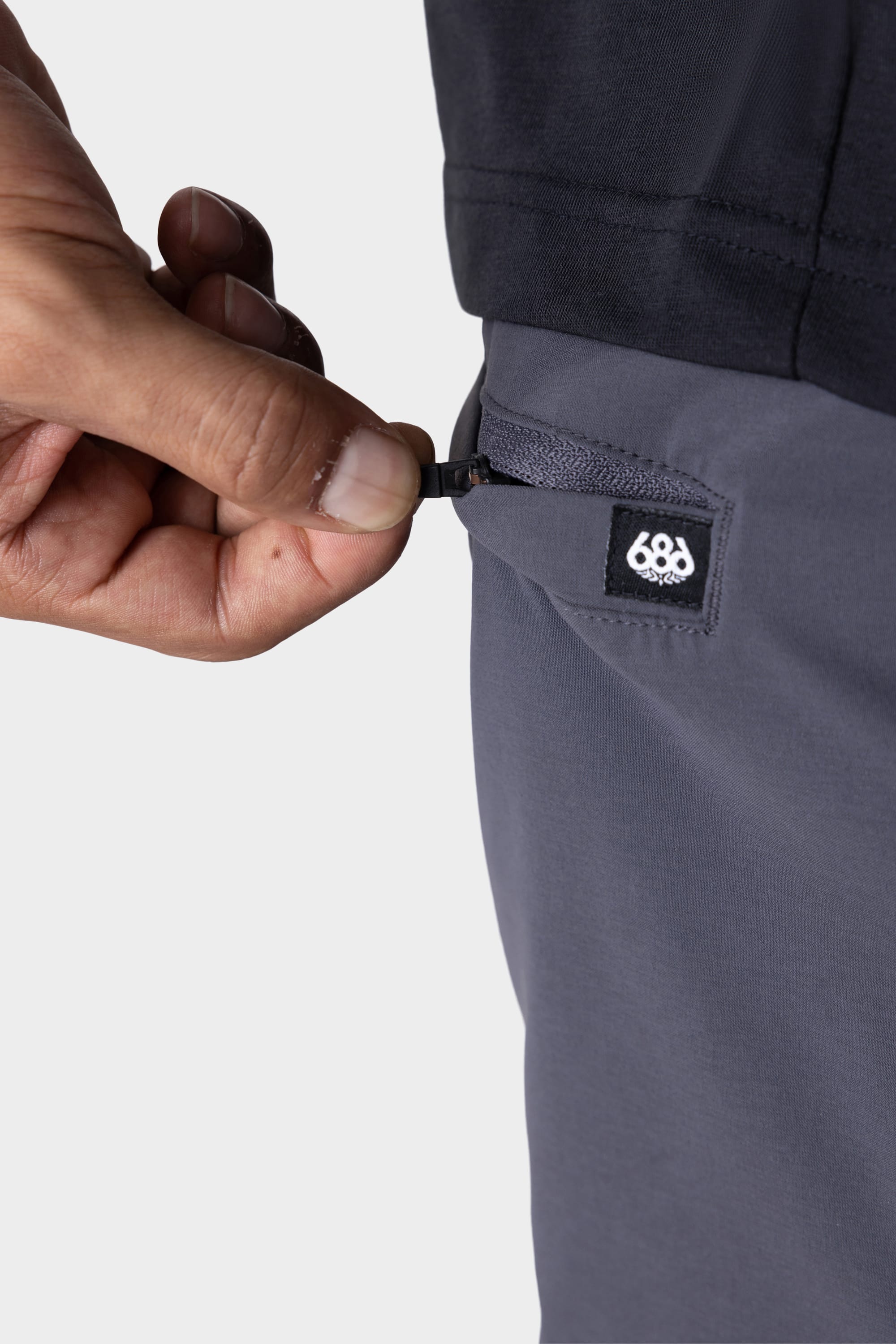 A close-up of a hand pulling the zipper on a small pocket of the 686 Men's Everywhere® Featherlight Chino Pant - Slim Fit, shown in gray with the 686 logo above the zipper—perfect for a summer wardrobe.