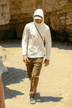 A person in a white hooded long-sleeve shirt, sunglasses, and 686 Men's Everywhere® Featherlight Chino Pant - Slim Fit walks with a crossbody strap on a sunny, sandy path—ideal for summer style.