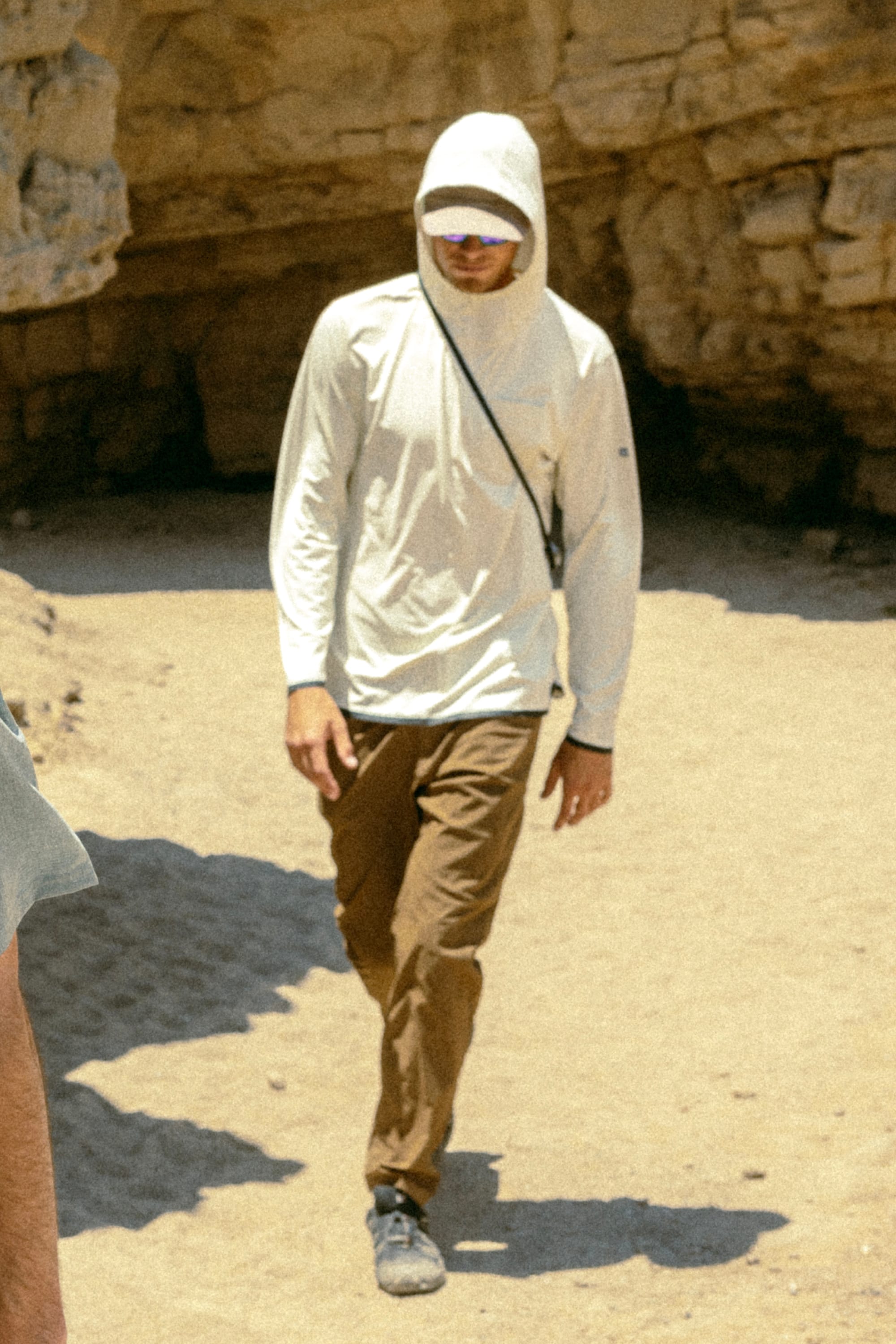 A person in a white hooded long-sleeve shirt, sunglasses, and 686 Men's Everywhere® Featherlight Chino Pant - Slim Fit walks with a crossbody strap on a sunny, sandy path—ideal for summer style.