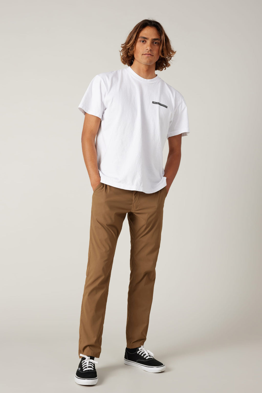 A young man with medium-length brown hair stands with his hands in his pockets, wearing a white T-shirt, black sneakers, and 686 Men's Everywhere® Featherlight Chino Pant - Slim Fit against a plain light background for a relaxed summer look.