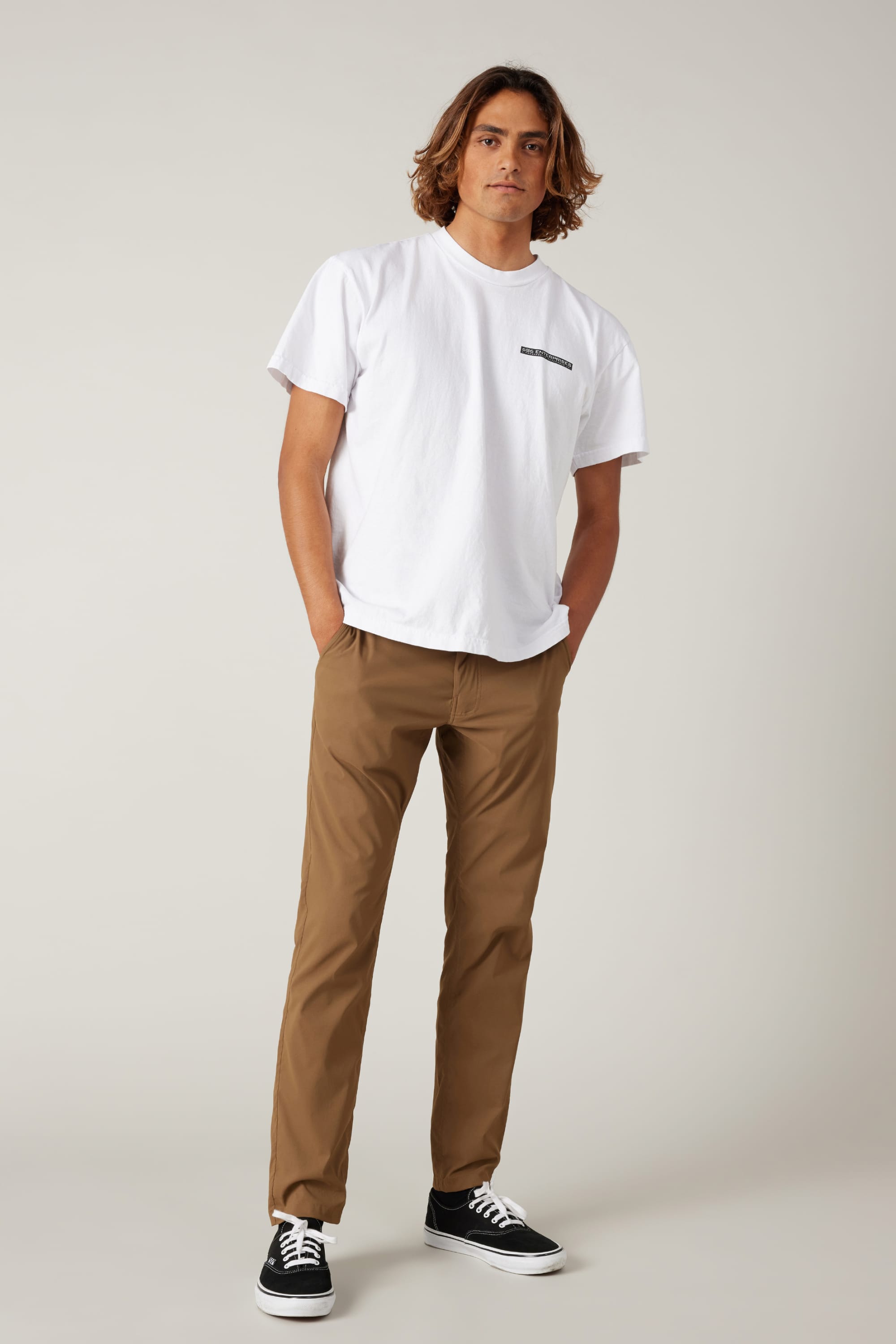 A young man with medium-length brown hair stands with his hands in his pockets, wearing a white T-shirt, black sneakers, and 686 Men's Everywhere® Featherlight Chino Pant - Slim Fit against a plain light background for a relaxed summer look.