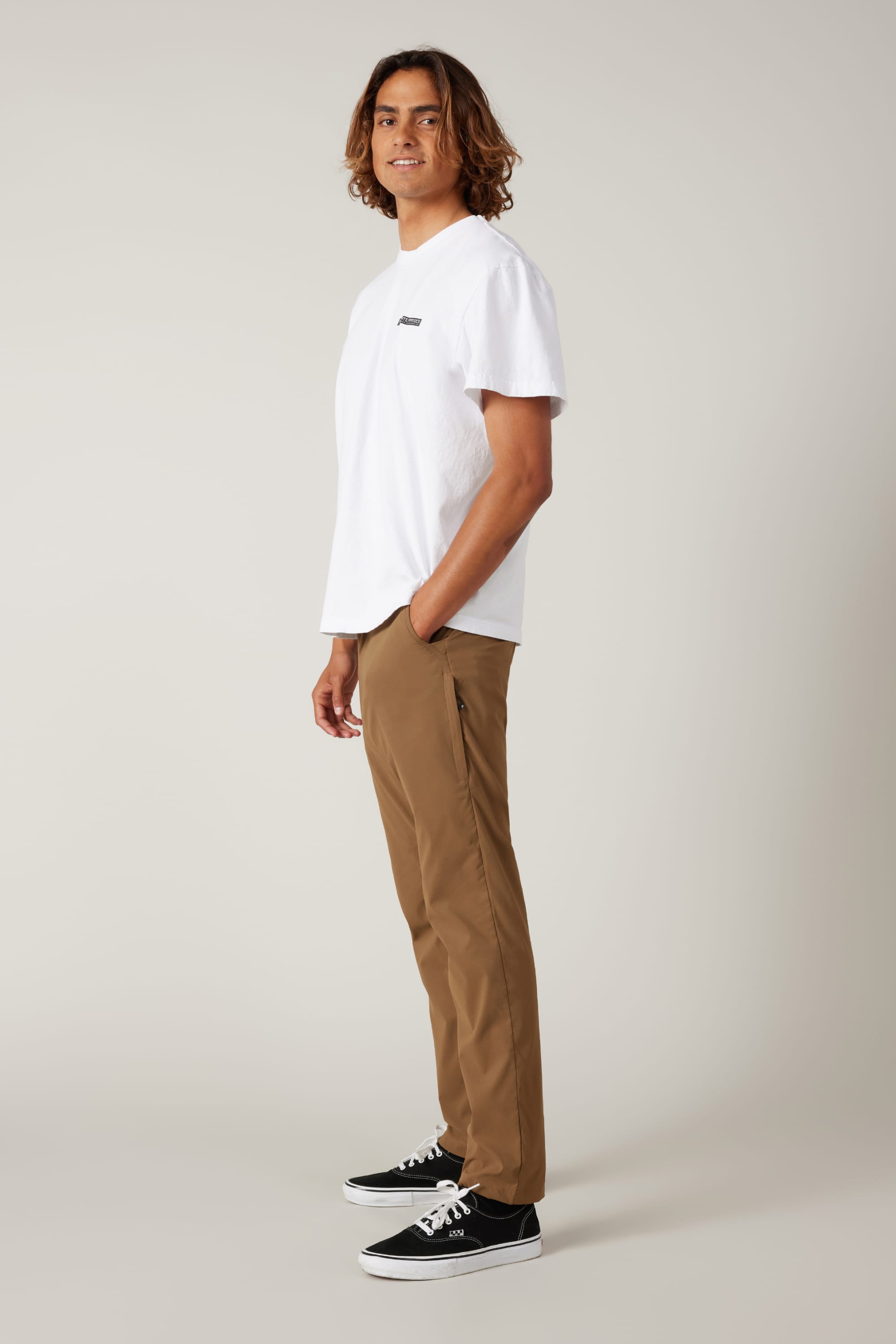 A young man with medium-length brown hair stands sideways, hands in pockets, wearing a white t-shirt, 686 Men's Everywhere® Featherlight Chino Pant - Slim Fit, and black sneakers—perfect for a fresh summer wardrobe.