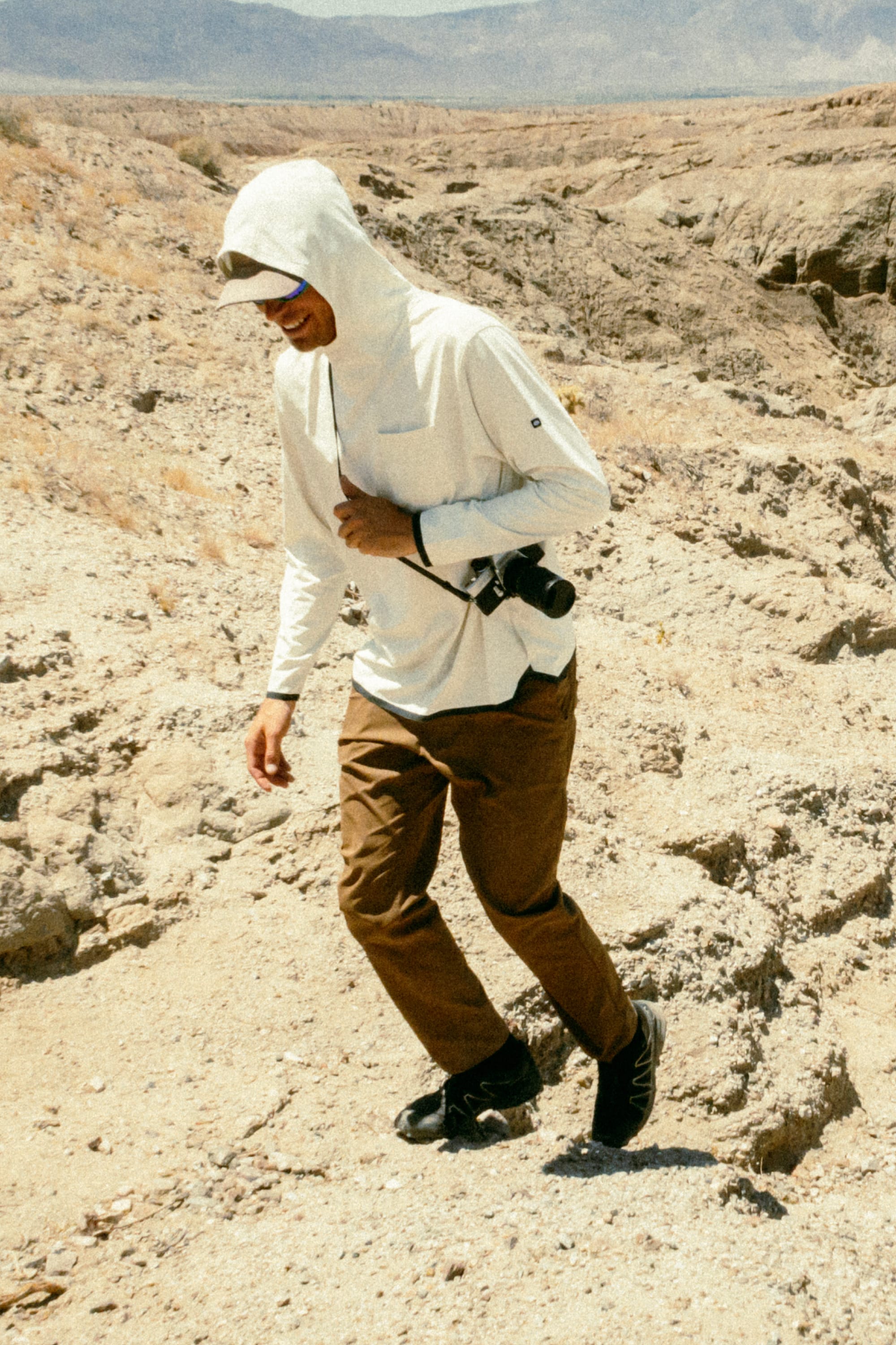 Wearing a breathable hooded shirt, 686 Men's Everywhere® Featherlight Chino Pant - Slim Fit, and sunglasses, a person walks outdoors in a dry, rocky desert landscape with a camera—ideal summer wardrobe from 686.