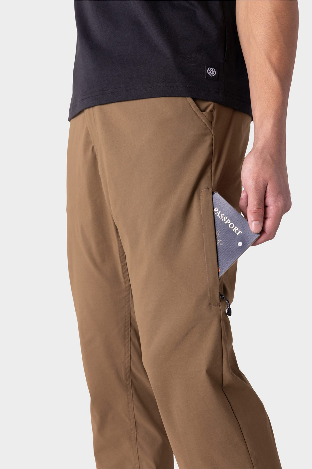 A person in 686 Men's Everywhere® Featherlight Chino Pant - Slim Fit and a black shirt places a passport into the pants' zippered side pocket. The plain, light background highlights this breathable summer essential from 686.