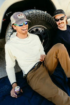Two men in sunglasses and summer attire relax by a truck tire. One, in 686 Men's Everywhere® Featherlight Chino Pant - Slim Fit, holds a canned drink and smiles. Both enjoy the moment together.